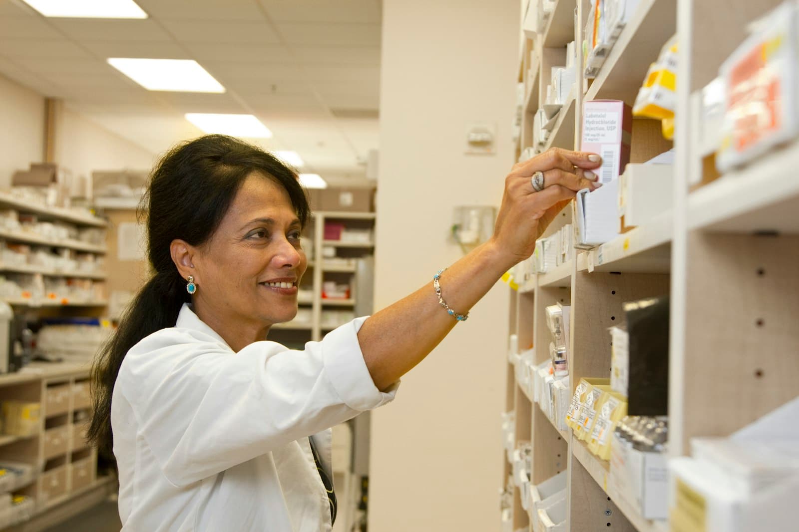 A smiling pharmacist reaching for medicine on a pharmacy shelf
